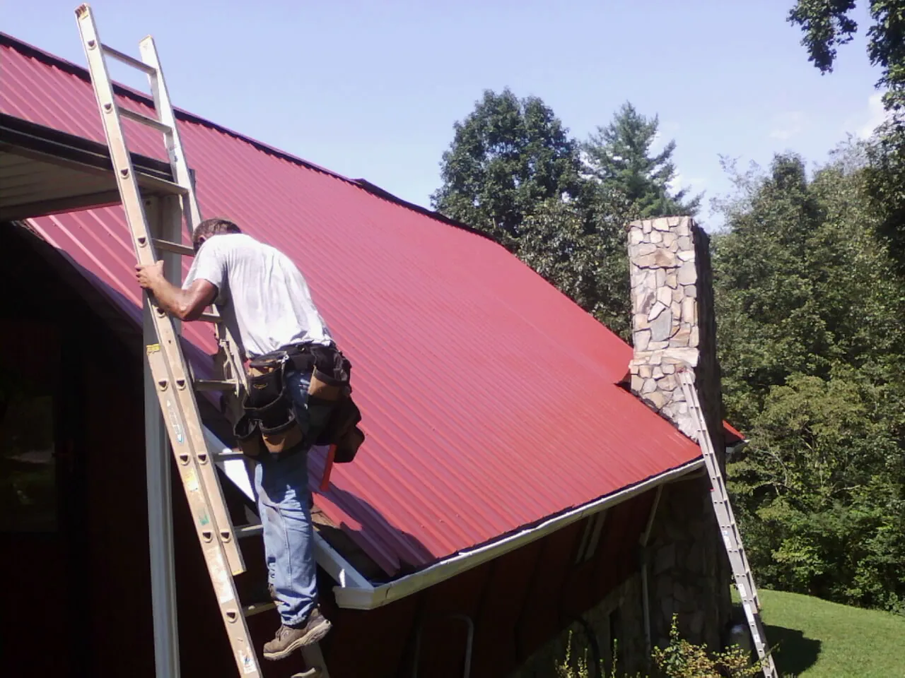 Roofer on a ladder beside a red metal roof and stone chimney — Soffit & Fascia Repair by Roofing Brothers in New Weston